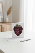 A white greeting card featuring a pressed flower art of a strawberry made from dark burgundy and purple dried flower petals with green leaves, shown with a white envelope on a light gray background.