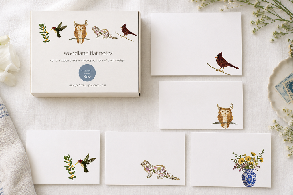 A flat-lay of a woodland-themed card and envelope set on a white fabric background. The set includes a box labeled 'woodland flat notes' with illustrated birds, a card, envelopes, and matching printed cards featuring drawings of an owl, a bird, a hummingbird, a floral animal, and a flower vase.