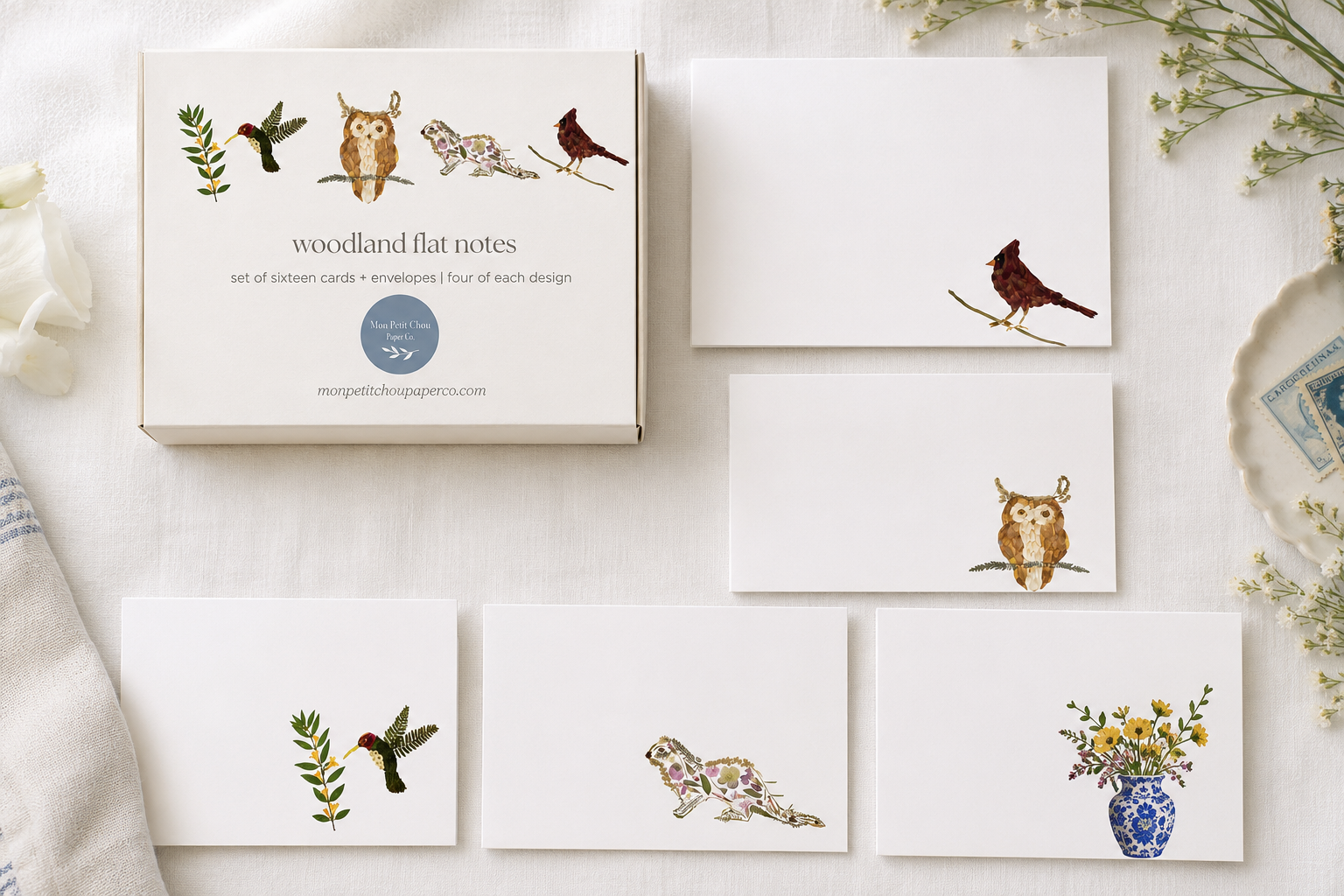 A flat-lay of a woodland-themed card and envelope set on a white fabric background. The set includes a box labeled 'woodland flat notes' with illustrated birds, a card, envelopes, and matching printed cards featuring drawings of an owl, a bird, a hummingbird, a floral animal, and a flower vase.