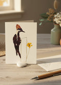 A pressed flat-lay art piece featuring a woodpecker crafted from dark brown and black leaves, a white chest detail, a reddish-brown crest, and a small yellow flower, set against a clean white background.